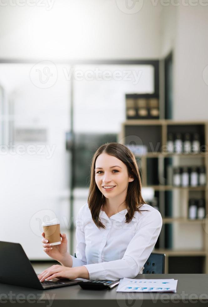 giovane bellissimo donna digitando su tavoletta e il computer portatile mentre seduta a il Lavorando di legno tavolo moderno ufficio foto