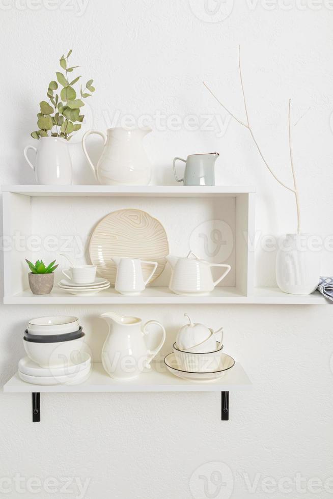 elegante bianca di legno scaffali su un' bianca strutturato parete con un' impostato di piatti di loro eco materiali. bellissimo design nel un' moderno cucina. foto