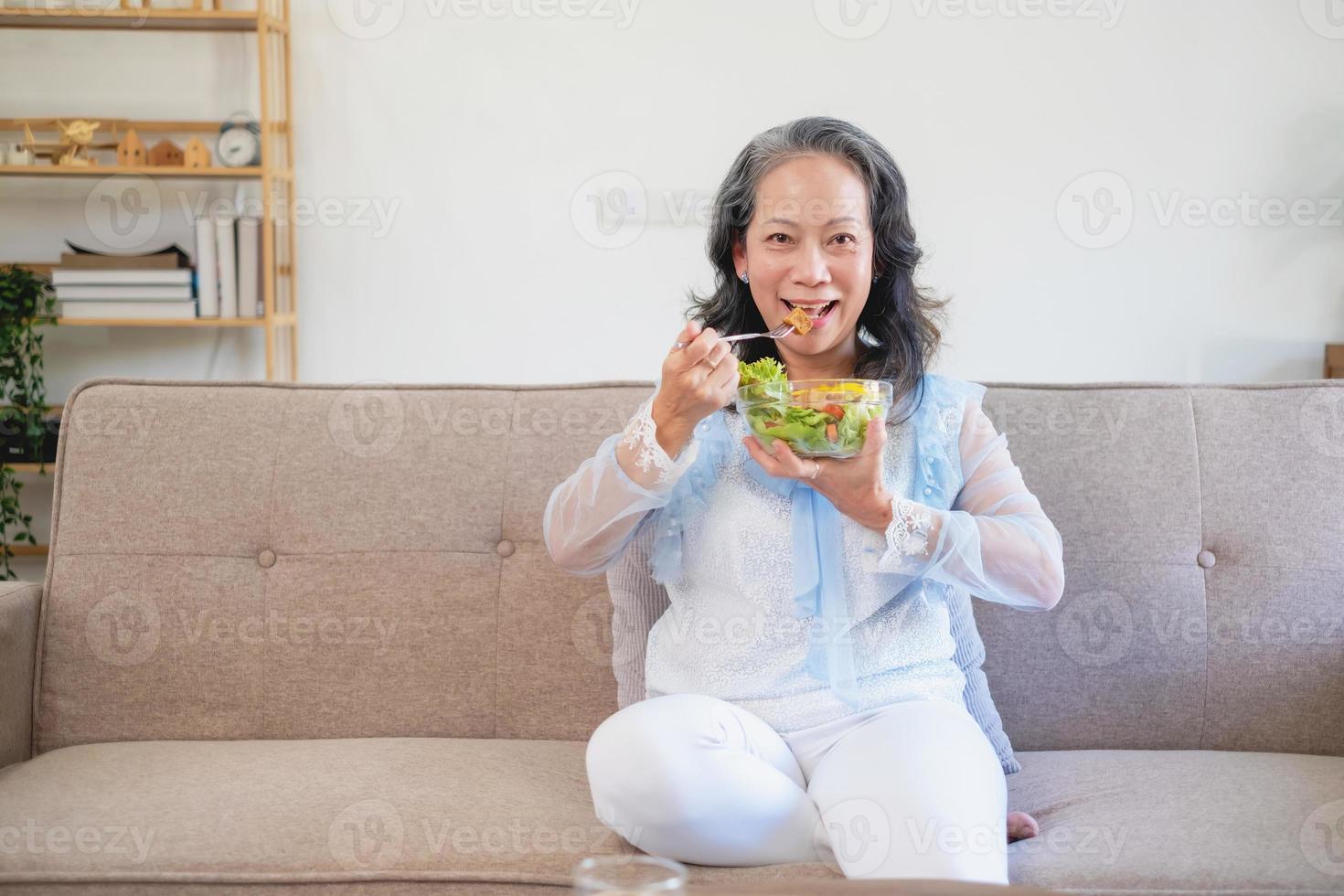 asiatico anziano donna seduta mangiare verdura insalata e salutare cibo e mangiare felicemente su il divano nel il Casa per un' salutare corpo. salutare cibo concetto foto