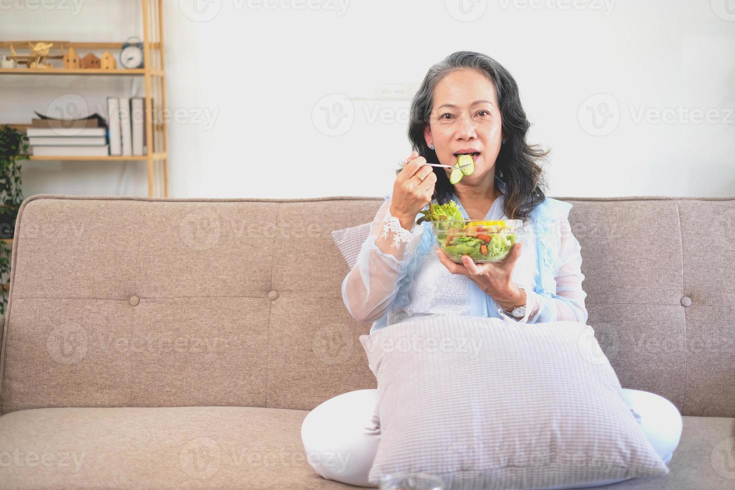asiatico anziano donna seduta mangiare verdura insalata e salutare cibo e mangiare felicemente su il divano nel il Casa per un' salutare corpo. salutare cibo concetto foto