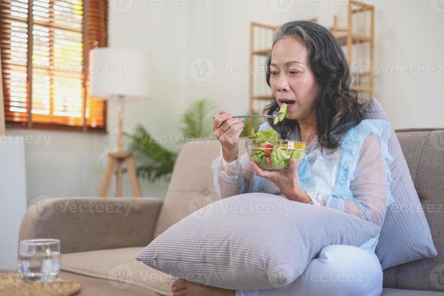 asiatico anziano donna seduta mangiare verdura insalata e salutare cibo e mangiare felicemente su il divano nel il Casa per un' salutare corpo. salutare cibo concetto foto