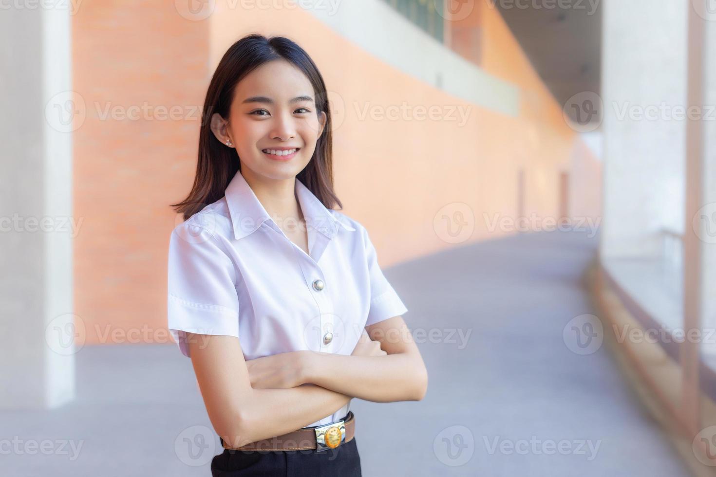 ritratto giovane asiatico bellissimo e bella tailandese ragazza alunno nel uniforme è sorridente guardare a telecamera e braccia attraversato per presente qualcosa con fiducia nel Università sfondo foto