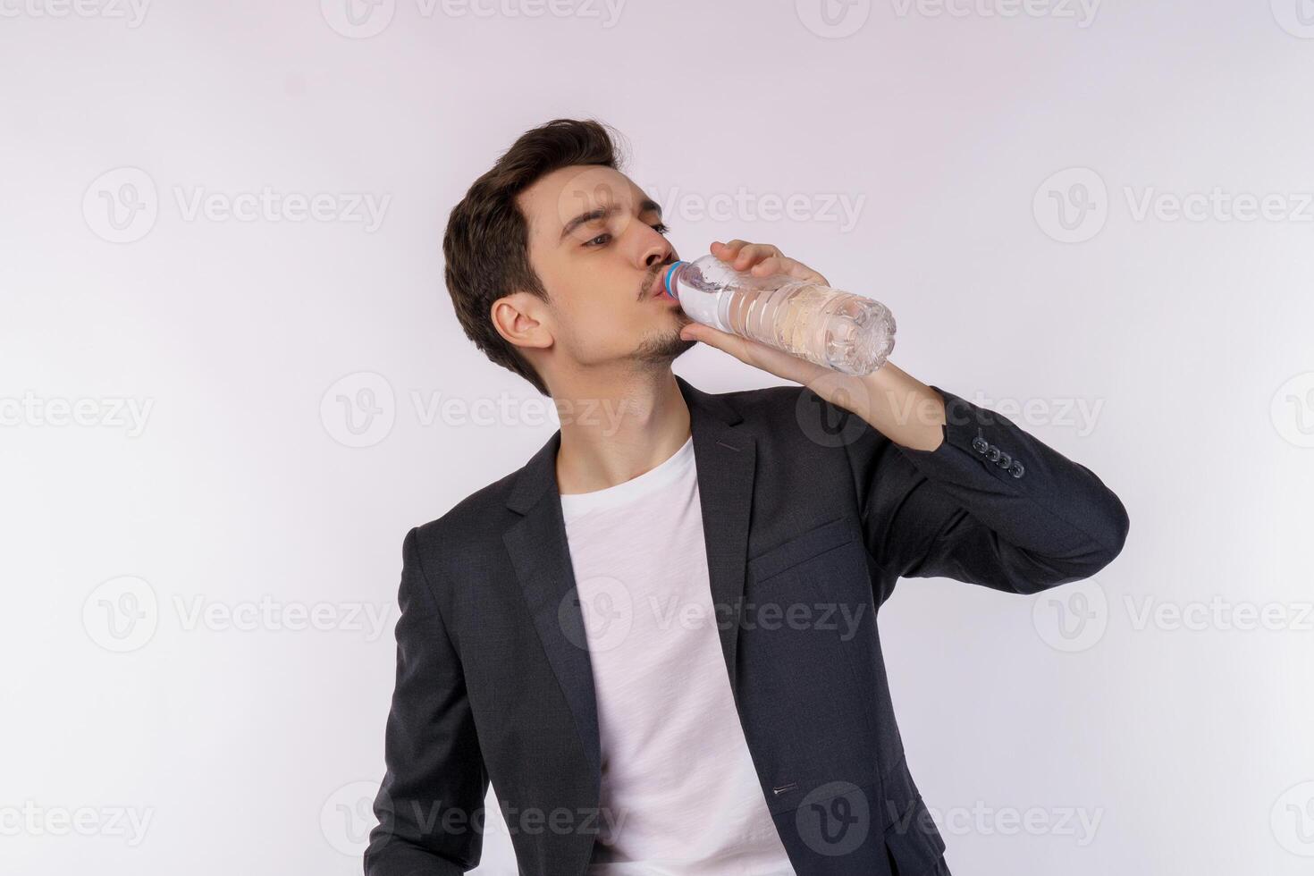 ritratto di contento giovane uomo potabile acqua a partire dal un' bottiglia e guardare a telecamera isolato al di sopra di bianca sfondo foto