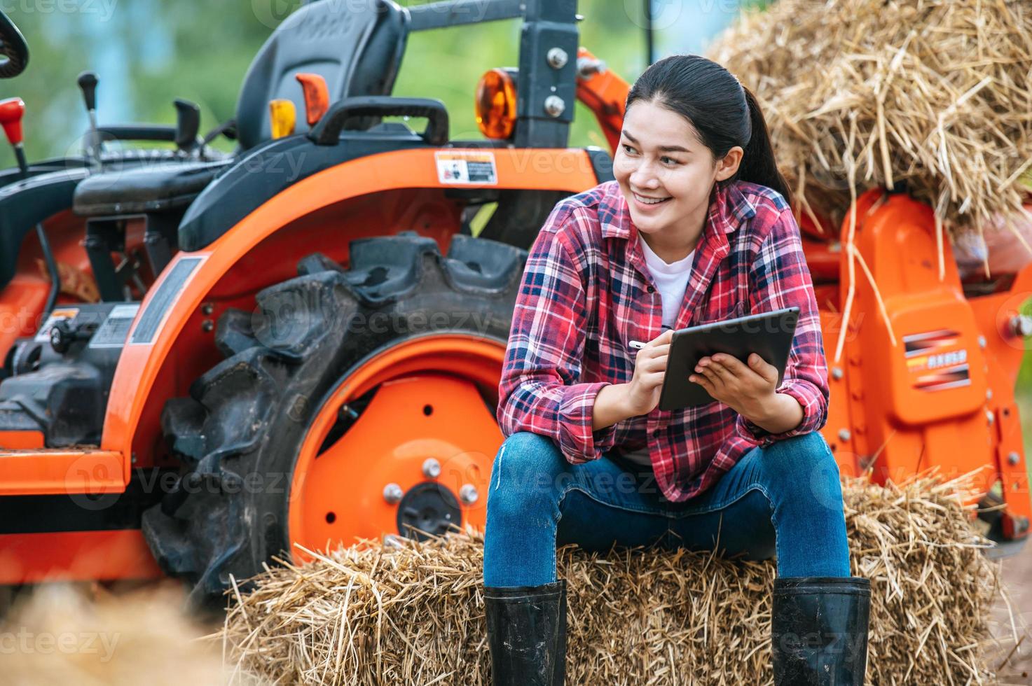 asiatico giovane femmina contadino seduta su balla di fieno con grande trattore macchina nel terreno agricolo. moderno tecnologie nel agricoltura gestione e agribusiness concetto. foto