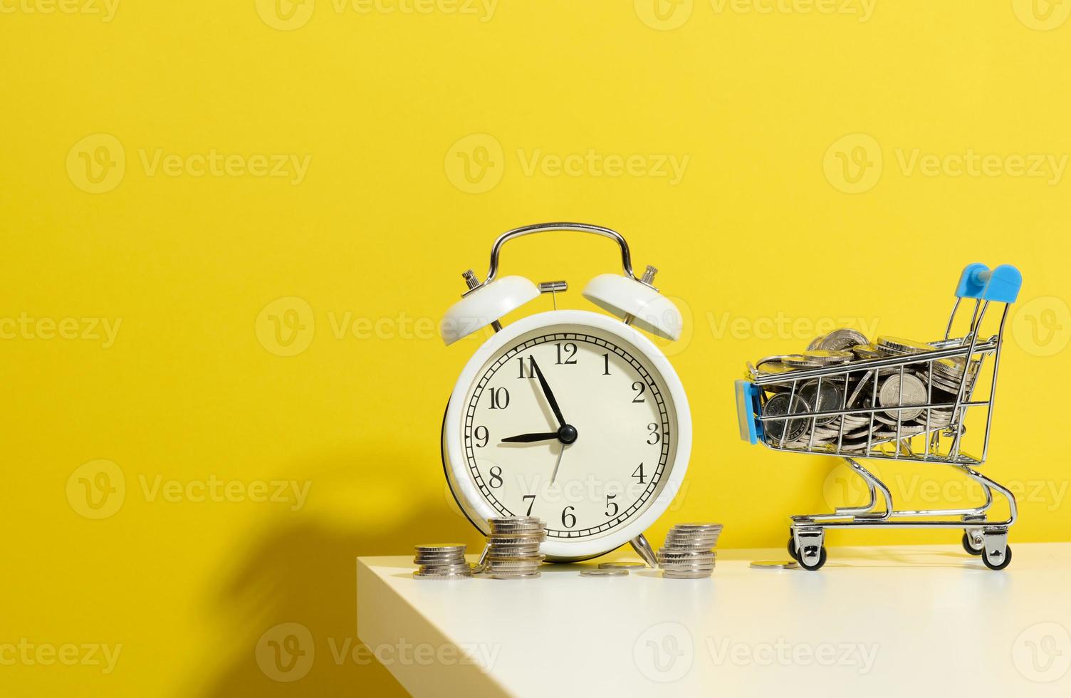 il giro allarme orologio, miniatura shopping carrello con monete su un' bianca tavolo. concetto tempo è i soldi, rifiuto di i soldi e povertà foto