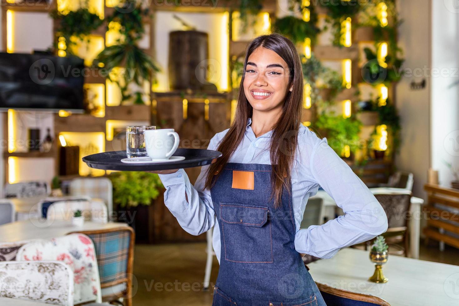 giovane donna Lavorando Tenere un' vassoio con caffè e bicchiere di acqua. bellissimo cameriera indossare grembiule. ritratto di un' sorridente cameriera Tenere vassoio di bevande nel ristorante foto