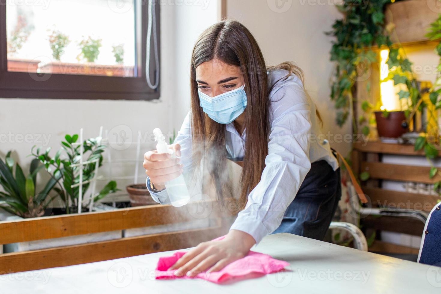 cameriera indossare protettivo viso maschera mentre disinfezione tavoli a ristorante o caffee per Il prossimo cliente. corona virus e piccolo attività commerciale è Aperto per opera concetto. foto