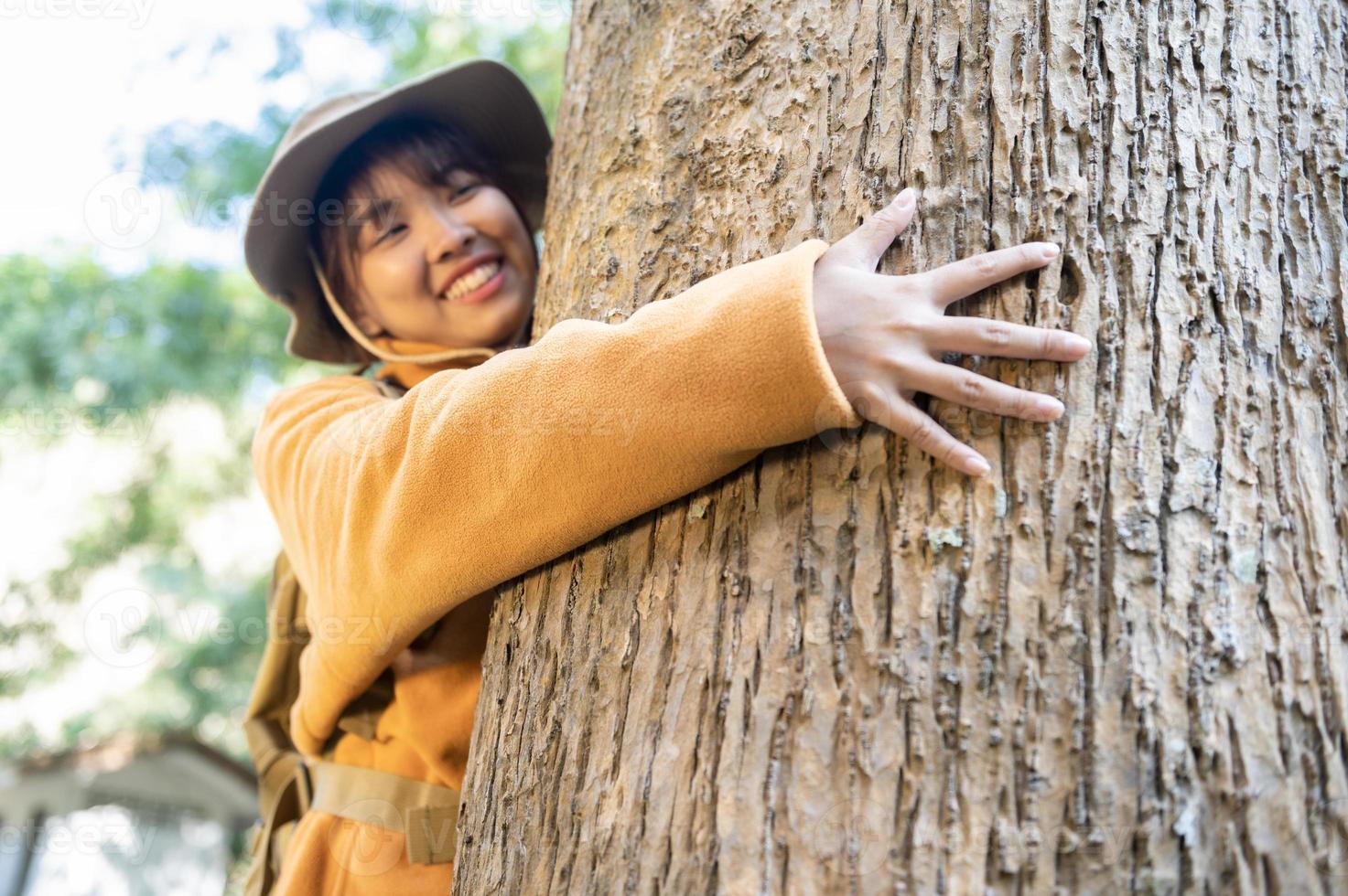 giovane turista donna nel un' giallo cappotto abbracciare un' albero nel il foresta di eco amore guardare su a il Treetops giovane asiatico donna l'esame un' grande ecologico albero foto