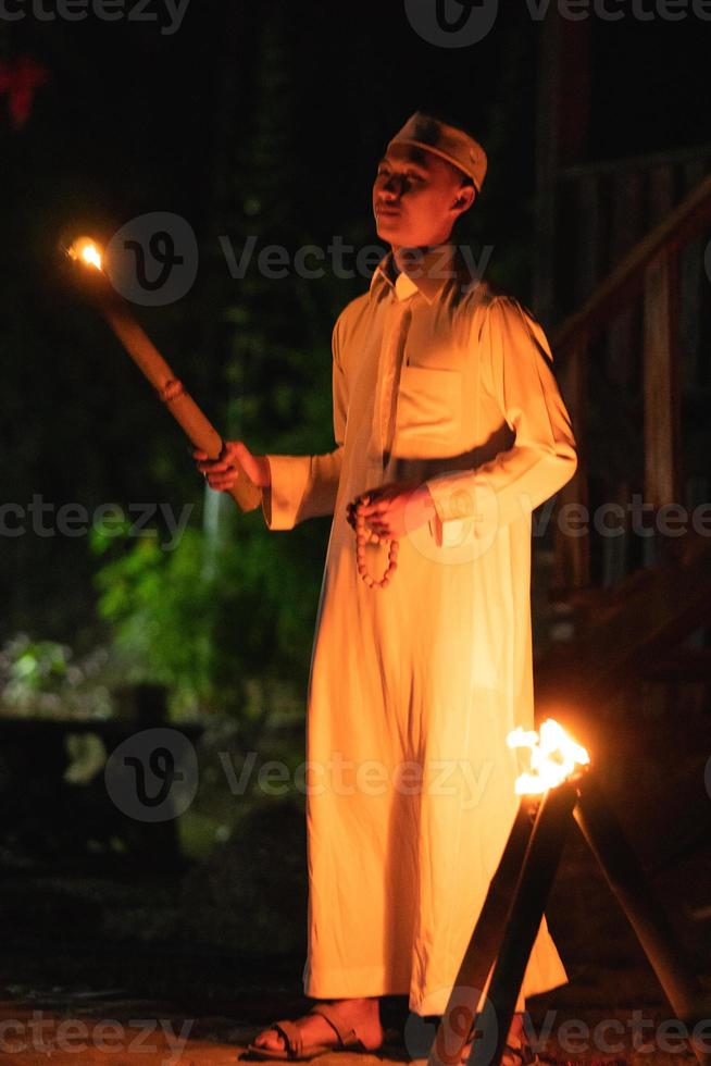 un' musulmano uomo in piedi con il fuoco torcia nel sua mano a il davanti di il villaggio foto