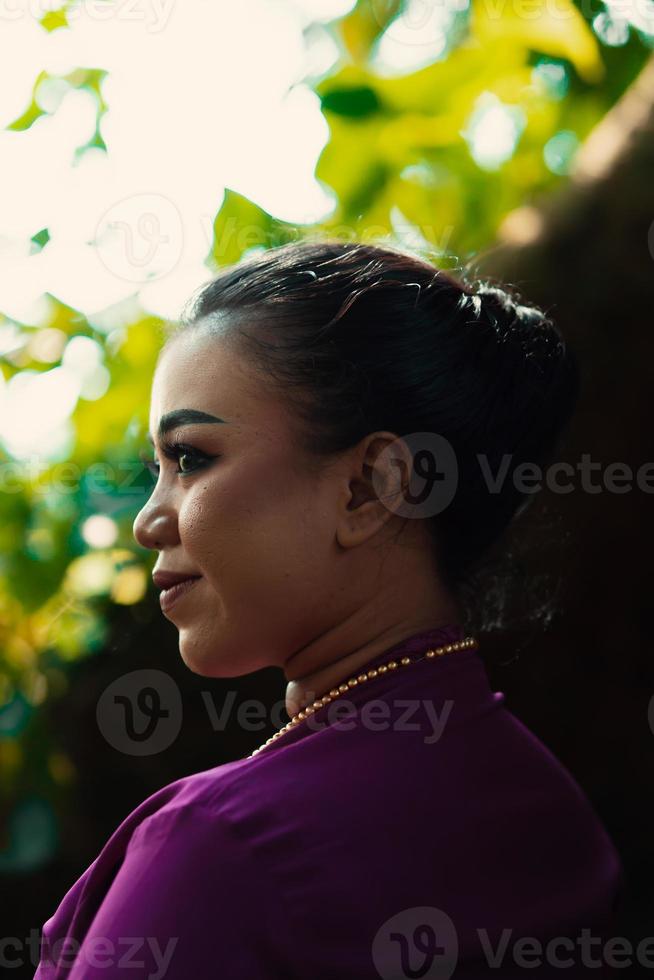 indonesiano donna sorridente nel un' tradizionale viola vestito mentre indossare trucco e d'oro gioielleria nel davanti di il grande albero foto