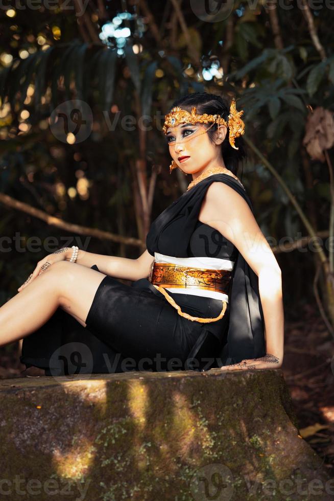 indonesiano donna seduta su il roccia nel un' nero danza costume mentre indossare un' d'oro corona e d'oro collana foto