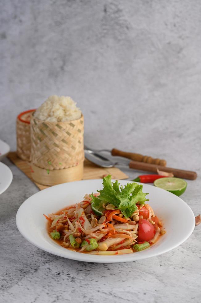 insalata di papaya tailandese su un piatto bianco con riso appiccicoso, peperoncino, cucchiaio e forchetta foto