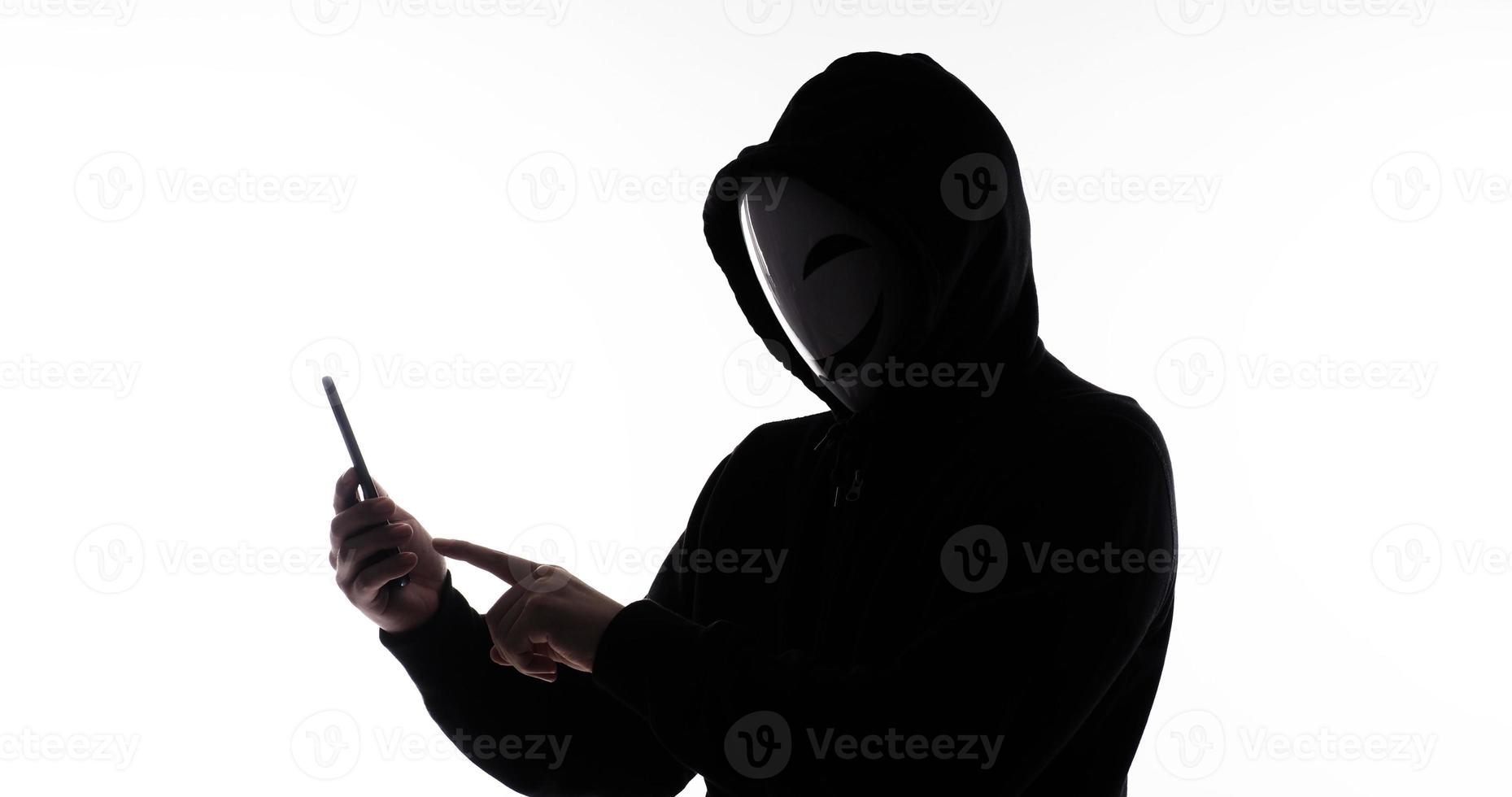 pirata anonimo e viso maschera con smartphone nel mano. uomo nel nero cappuccio camicia Tenere e utilizzando mobile Telefono su bianca sfondo. rappresentare informatica crimine dati pirateria o furto personale dati concetto foto