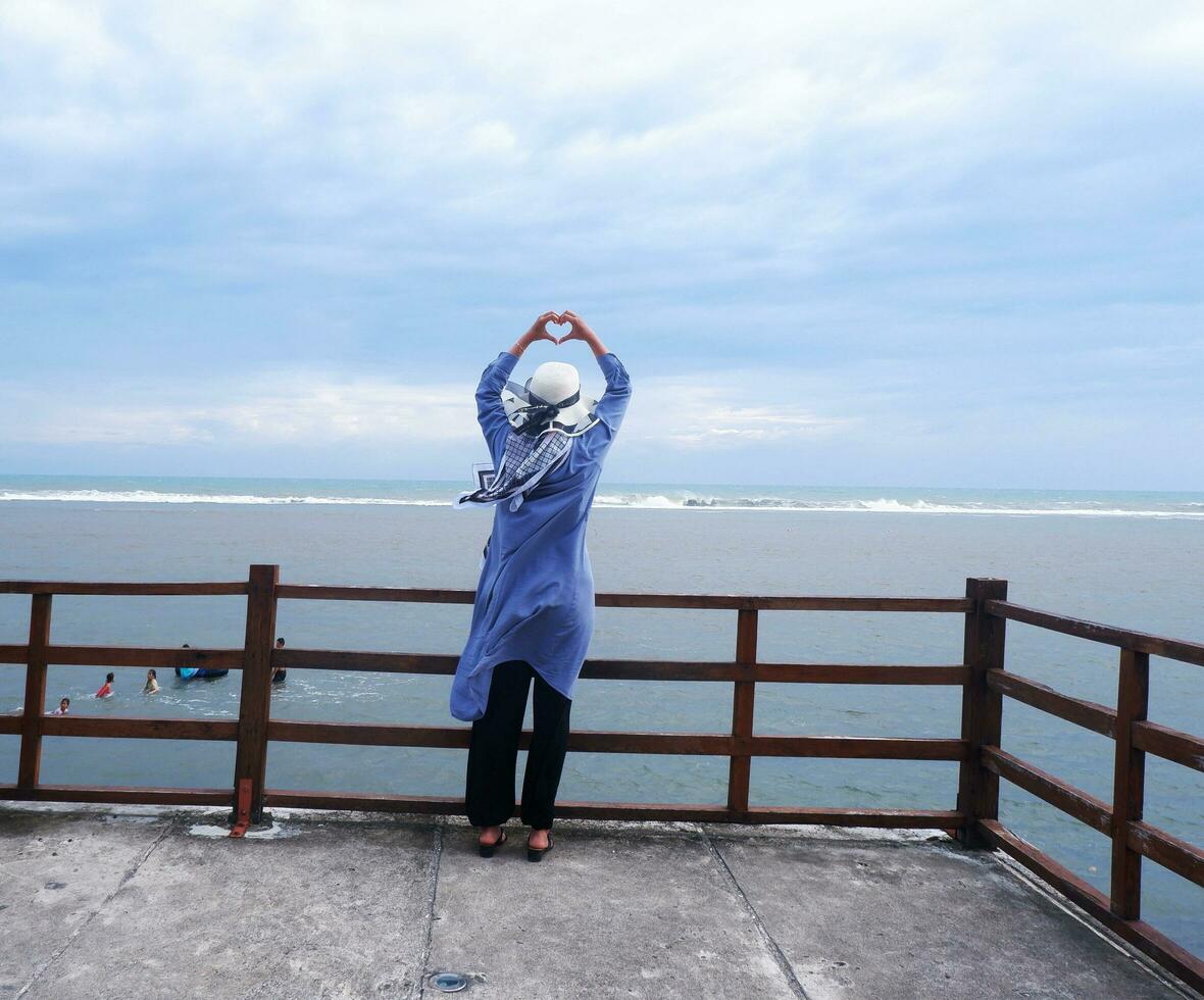 indietro di il donna nel il cappello su il tropicale spiaggia chi era guardare su a il cielo e il mare mentre sua mani formato amore a partire dal il ponte. spiaggia Visualizza foto