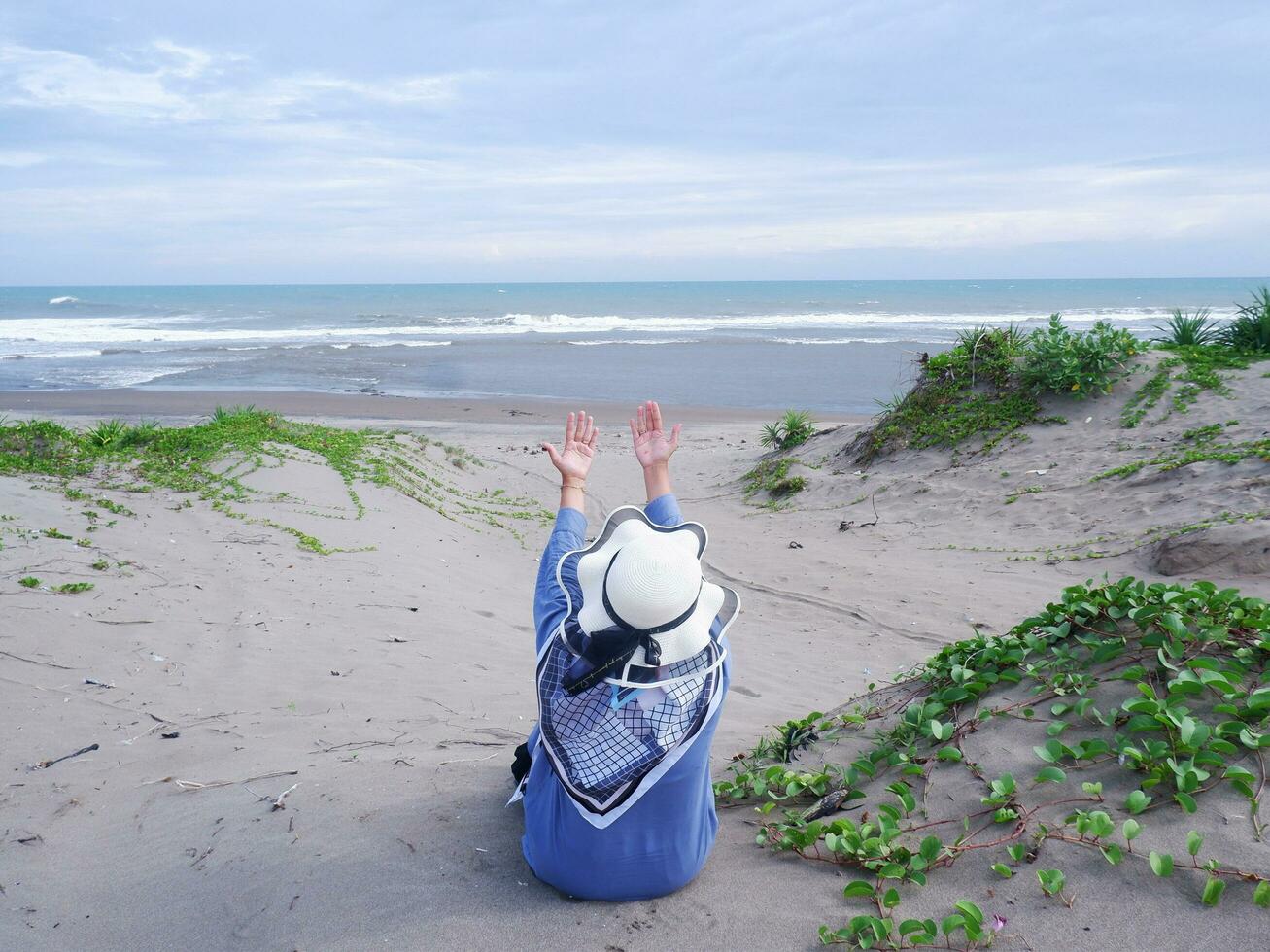 indietro di donna nel il cappello su il tropicale spiaggia chi era seduta su il sabbia e guardare a il cielo e il mare, mentre diffusione sua braccia . contento donne.spiaggia Visualizza foto