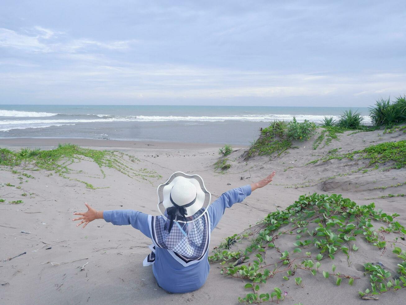 indietro di donna nel il cappello su il tropicale spiaggia chi era seduta su il sabbia e guardare a il cielo e il mare, mentre diffusione sua braccia . contento donne.spiaggia Visualizza foto