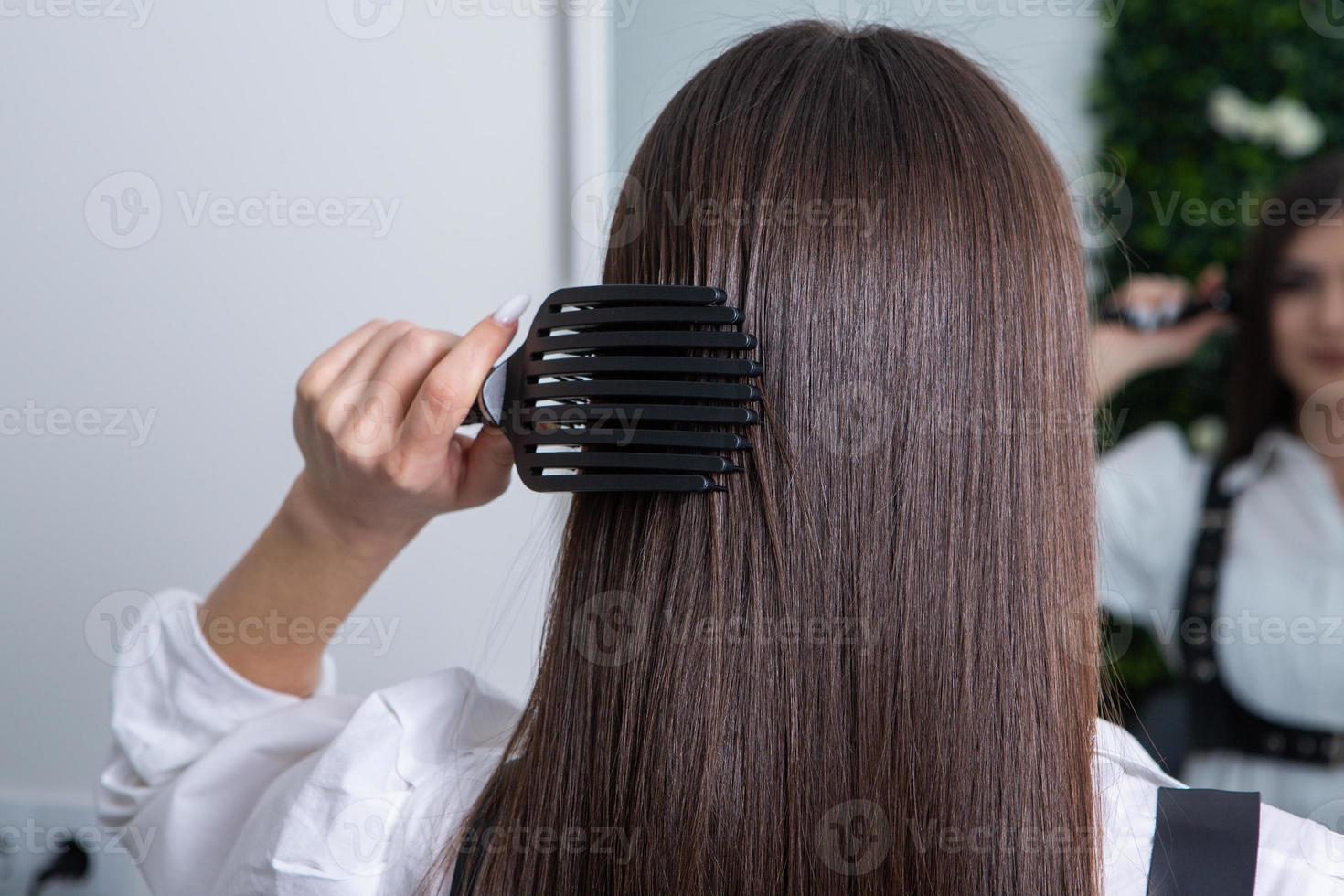 giovane donna pettinatura sua lungo buio capelli con un' pettine nel un' bellezza salone. un' dritto salutare brunetta capelli quello ha subito il capelli raddrizzatura procedura. foto