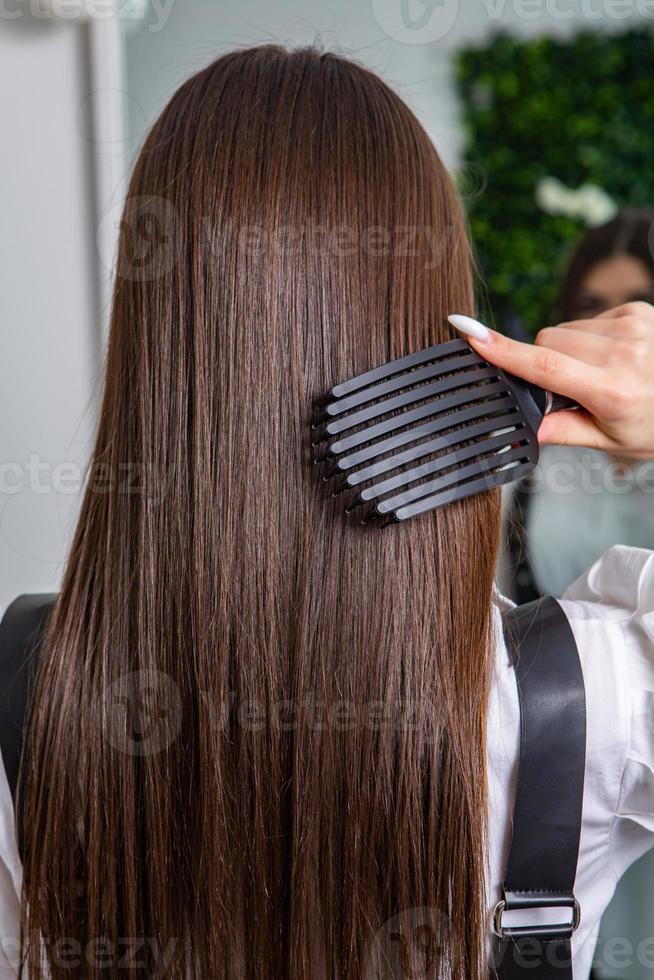 giovane donna pettinatura sua lungo buio capelli con un' pettine nel un' bellezza salone. un' dritto salutare brunetta capelli quello ha subito il capelli raddrizzatura procedura. foto