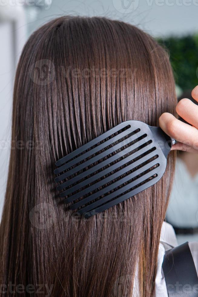 giovane donna pettinatura sua lungo buio capelli con un' pettine nel un' bellezza salone. un' dritto salutare brunetta capelli quello ha subito il capelli raddrizzatura procedura. foto