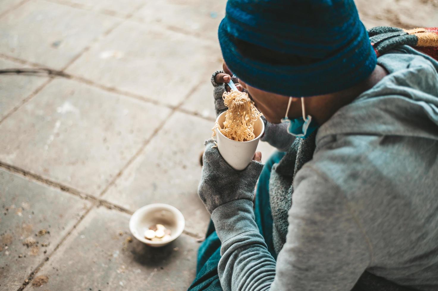 un senzatetto siede avvolto in un panno mangiando spaghetti foto
