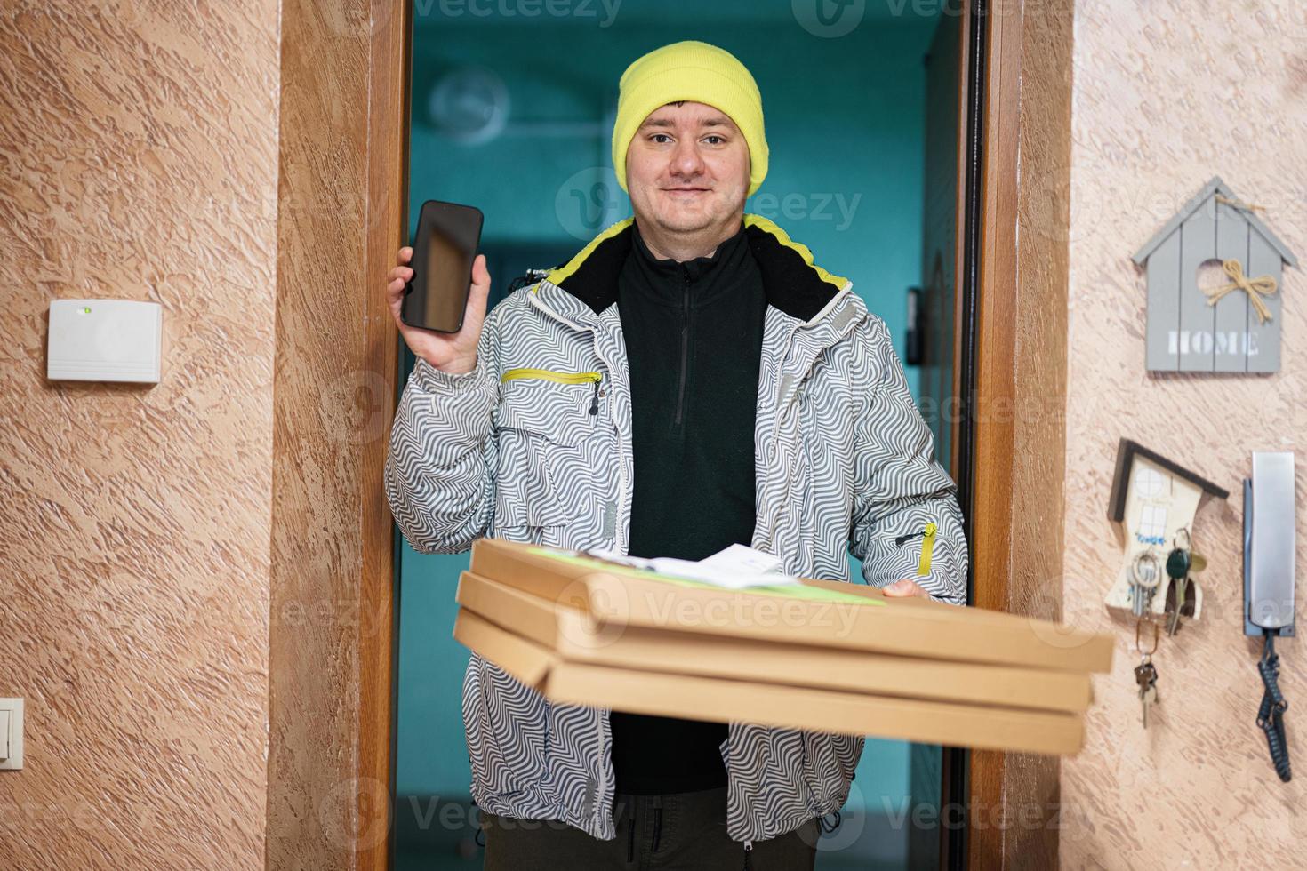 consegna uomo con Pizza cartone scatole. Corriere nel verde colore cappello con mobile Telefono in piedi contro porta di Residenziale Casa foto