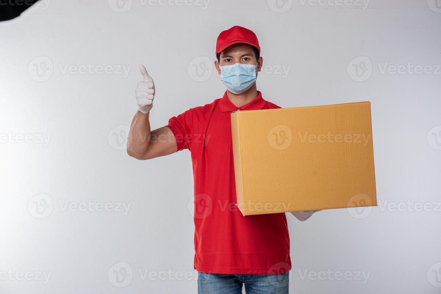 Immagine di un' consapevole giovane consegna uomo nel rosso berretto vuoto maglietta uniforme viso maschera guanti in piedi con vuoto Marrone cartone scatola isolato su leggero grigio sfondo studio foto