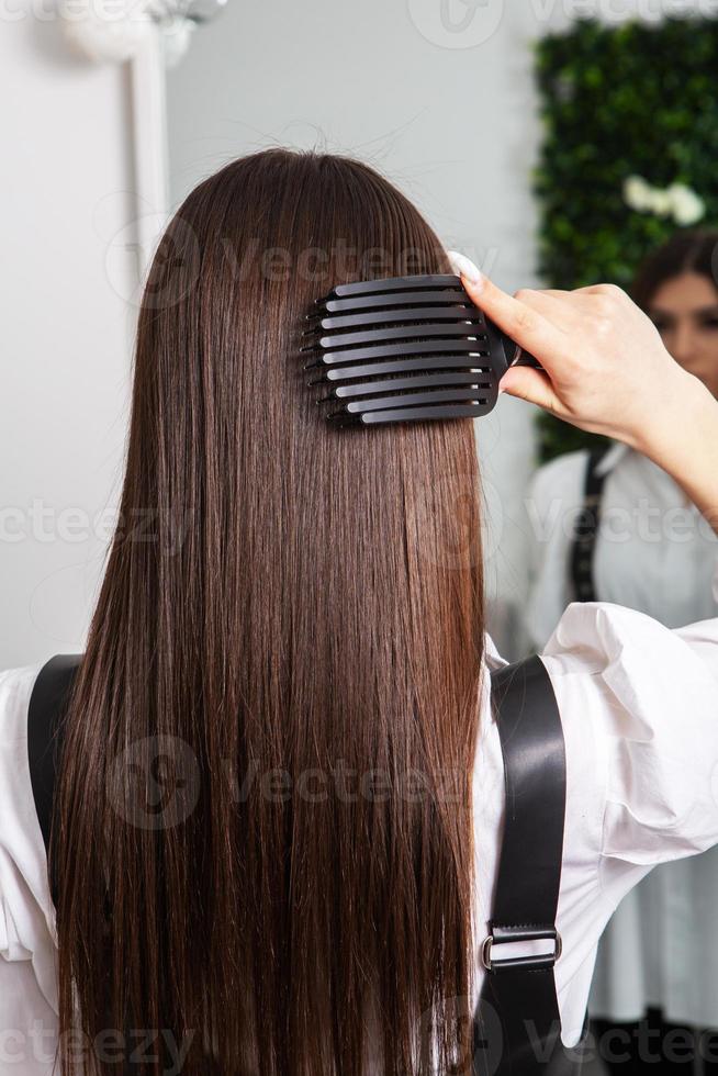 giovane donna pettinatura sua lungo buio capelli con un' pettine nel un' bellezza salone. un' dritto salutare brunetta capelli quello ha subito il capelli raddrizzatura procedura. foto