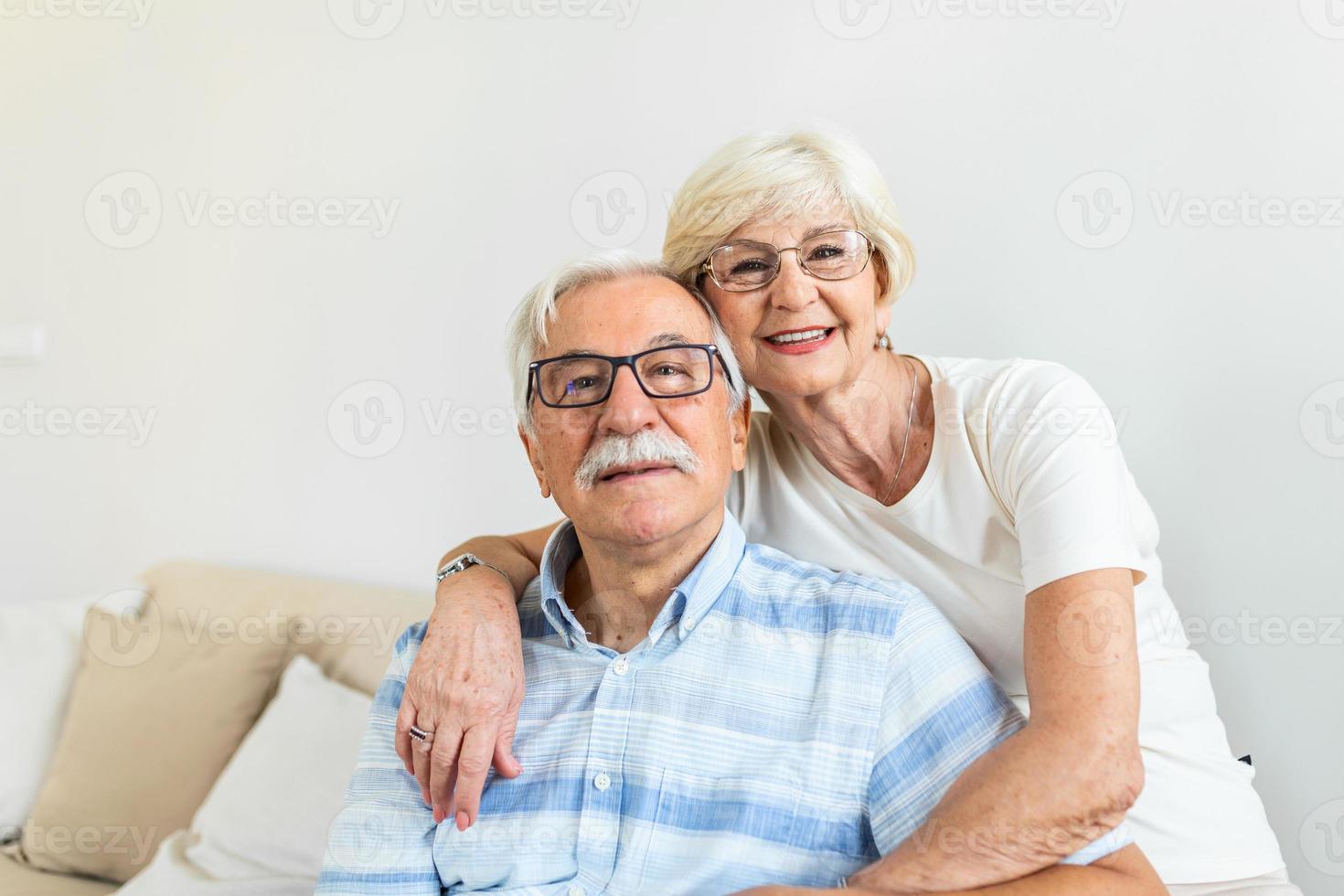 ritratto amorevole più vecchio moglie abbracciare marito seduta su accogliente divano. contento anziano maturo coppia smilling e guardare a telecamera, in posa per famiglia foto a casa. anziano coppia sensazione contento