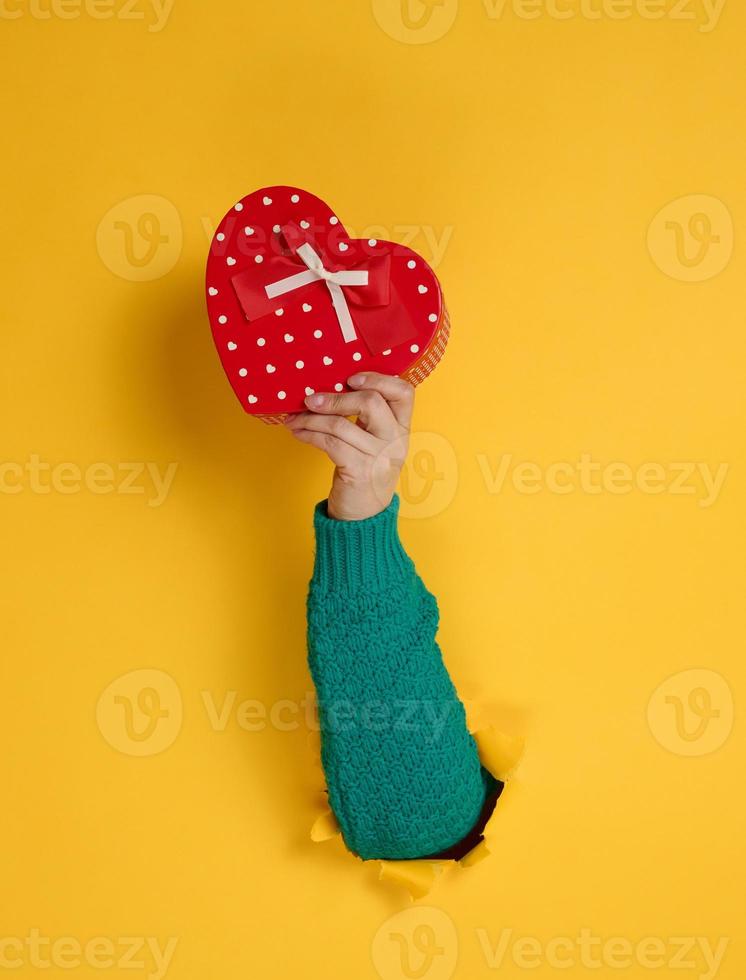 femmina mano detiene un' rosso scatola con un' regalo su un' giallo sfondo, parte di il corpo bastoni su di un' strappato buco nel un' carta sfondo. congratulazioni, vacanza sorpresa foto