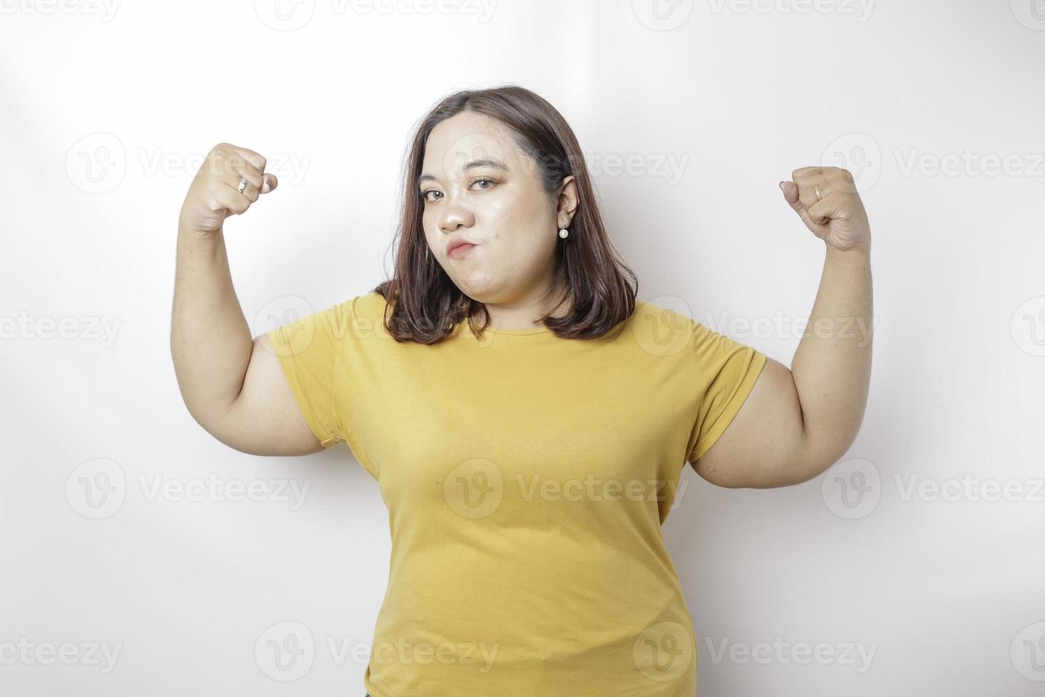 eccitato asiatico grande dimensioni donna indossare un' giallo maglietta mostrando forte gesto di sollevamento sua braccia e muscoli sorridente con orgoglio foto
