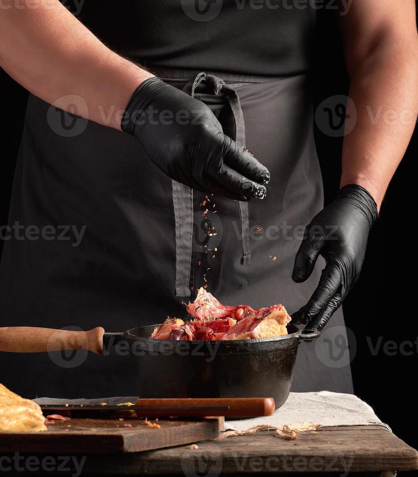 capocuoco nel nero uniforme e latice guanti stagionatura crudo pollo carne nel un' nero ghisa frittura padella foto