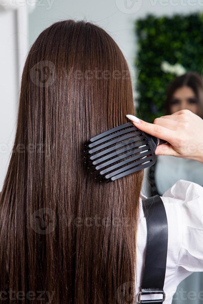 giovane donna pettinatura sua lungo buio capelli con un' pettine nel un' bellezza salone. un' dritto salutare brunetta capelli quello ha subito il capelli raddrizzatura procedura. foto