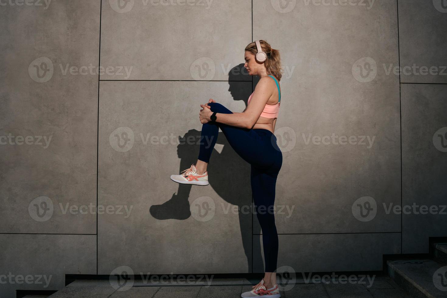 un' giovane ragazza nel cuffie sta vicino un' grigio parete tratti sua muscoli foto