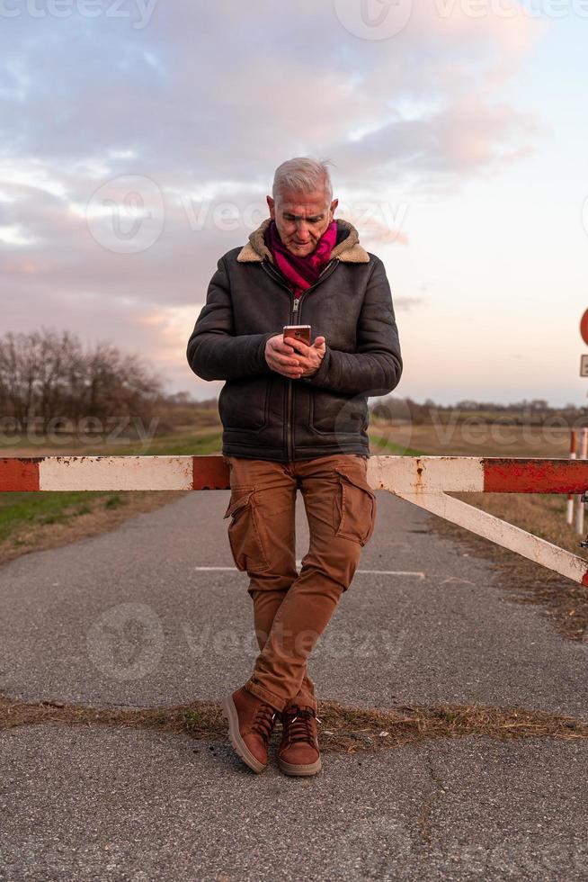 mezzo anziano uomo indossare inverno Abiti utilizzando smartphone nel un' nazione strada foto