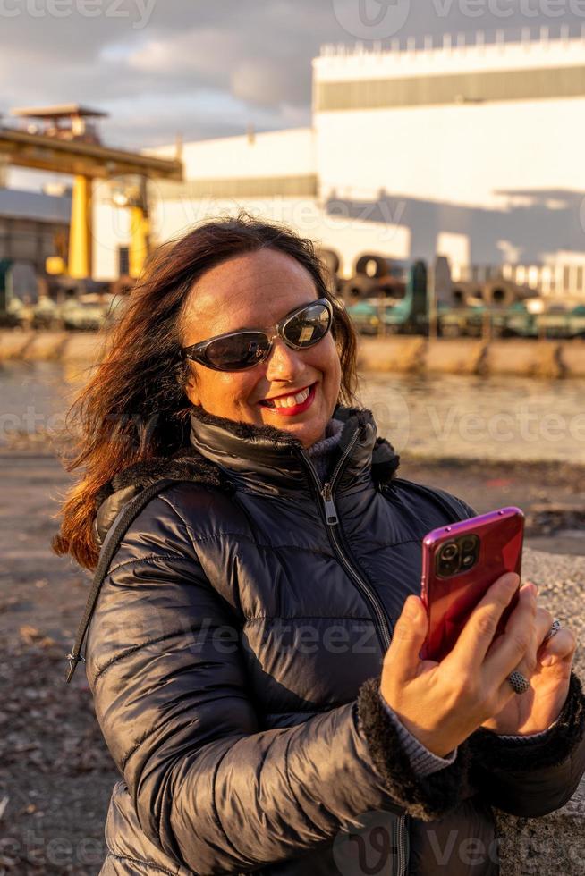 mezzo anziano donna indossare inverno Abiti utilizzando smartphone nel davanti di un' fabbrica foto