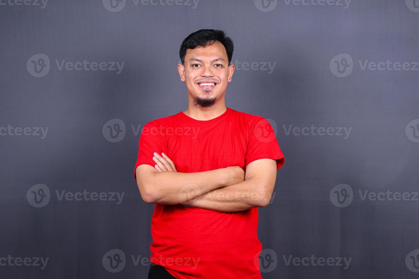 ritratto di attraente asiatico uomo nel rosso maglietta in piedi con attraversato braccia isolato su grigio sfondo foto