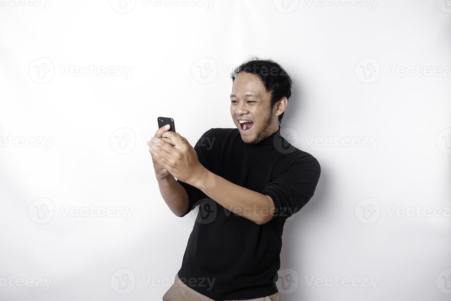 un' giovane asiatico uomo con un' contento riuscito espressione indossare nero camicia e Tenere il suo Telefono, isolato di bianca sfondo foto