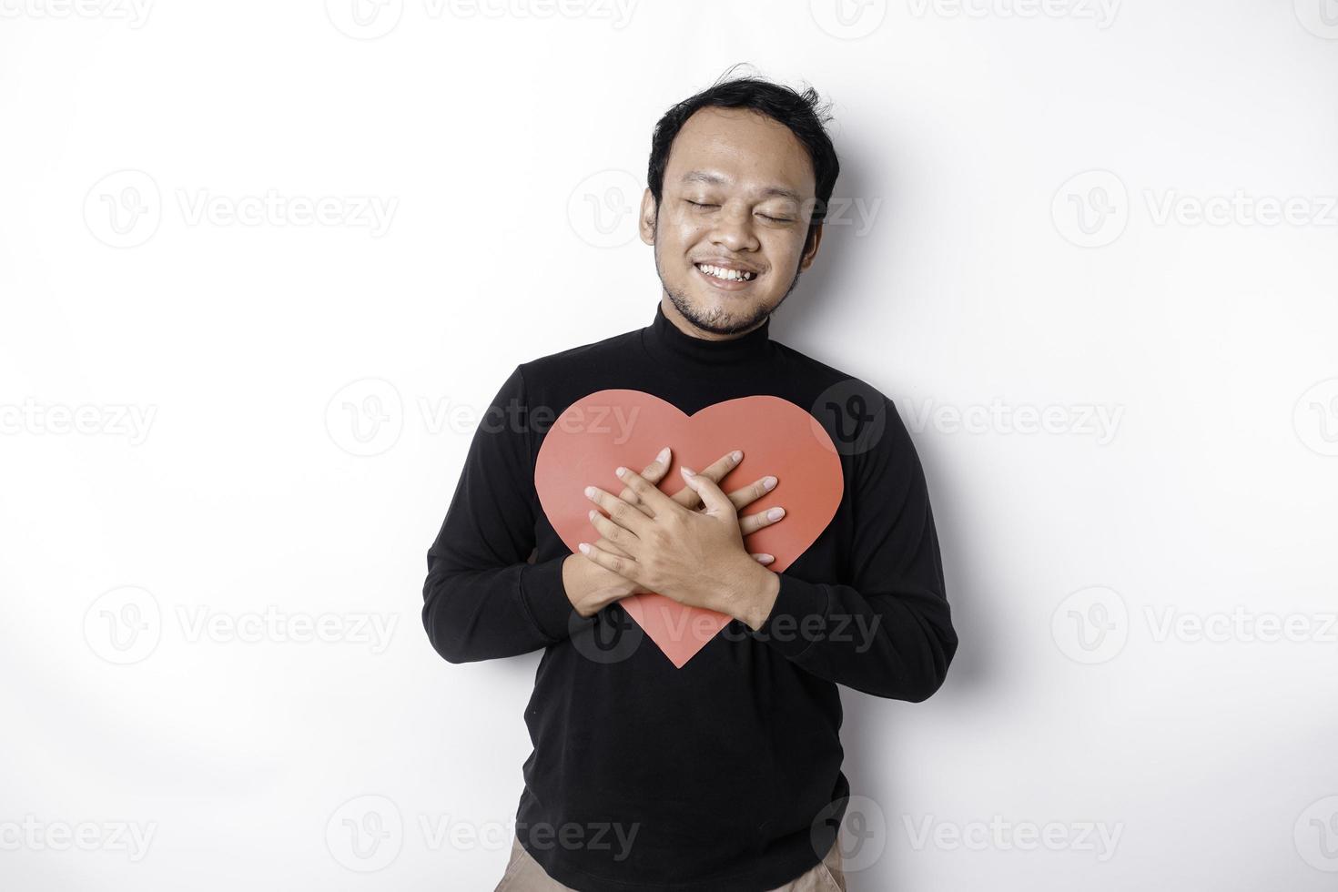 un' ritratto di un' contento asiatico uomo indossare un' nero camicia, Tenere un' rosso a forma di cuore carta isolato di bianca sfondo foto