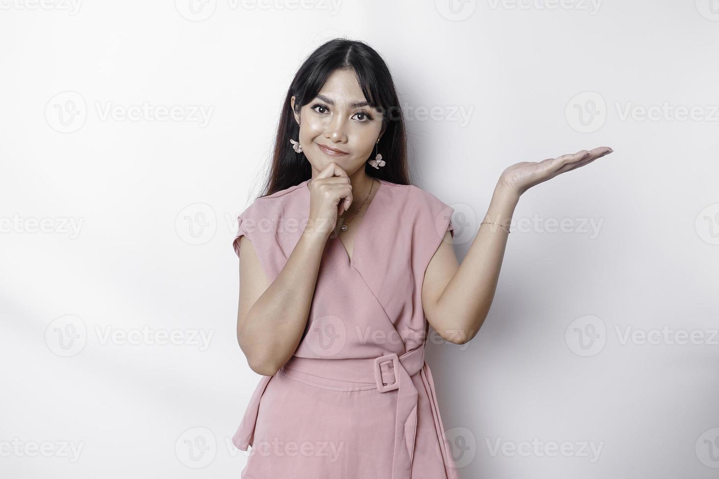 un' riflessivo giovane donna vestito nel rosa e Tenere sua mento mentre puntamento copia spazio accanto suo, isolato di bianca sfondo foto