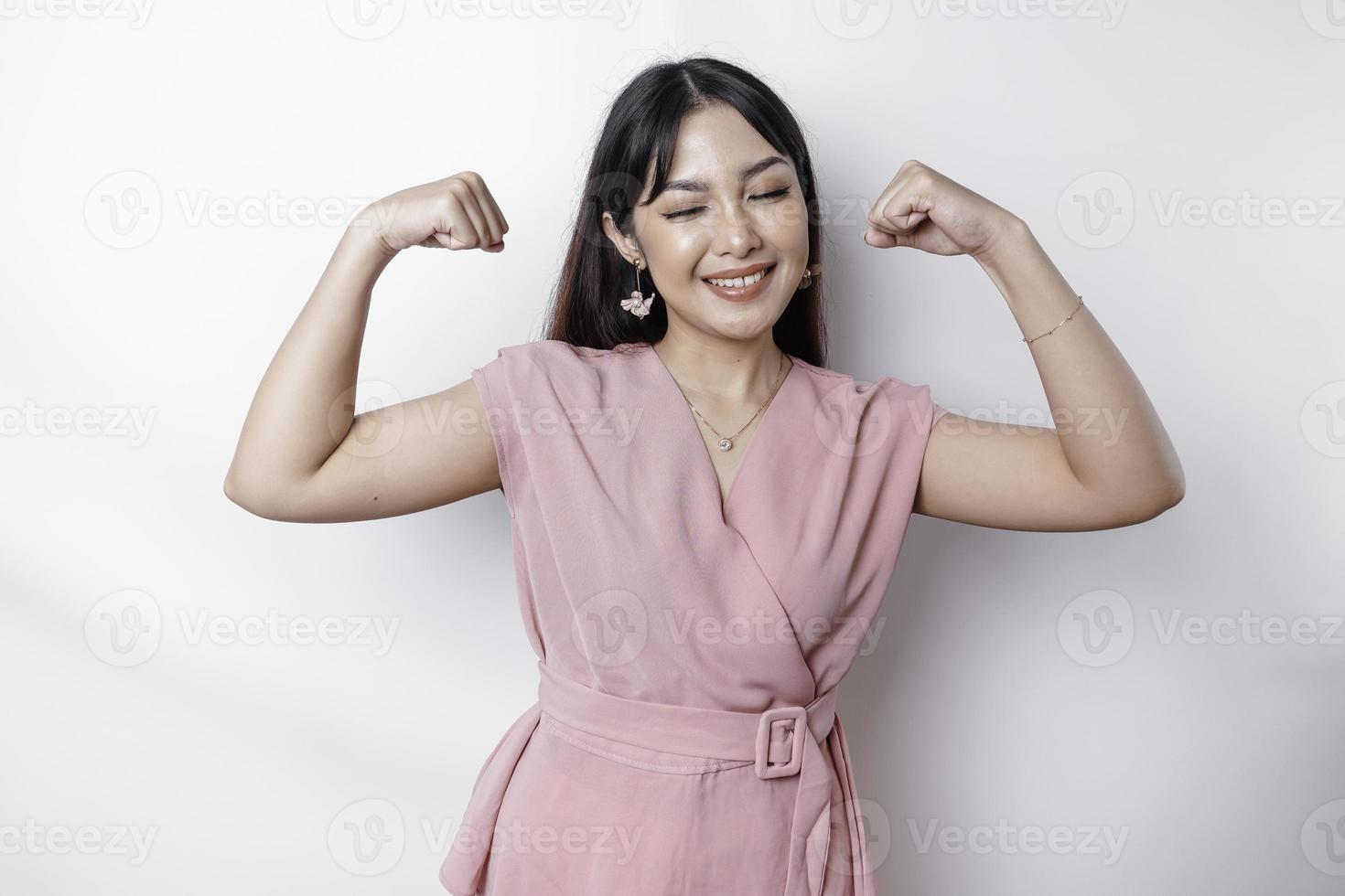 eccitato asiatico donna indossare un' rosa camicetta mostrando forte gesto di sollevamento sua braccia e muscoli sorridente con orgoglio foto