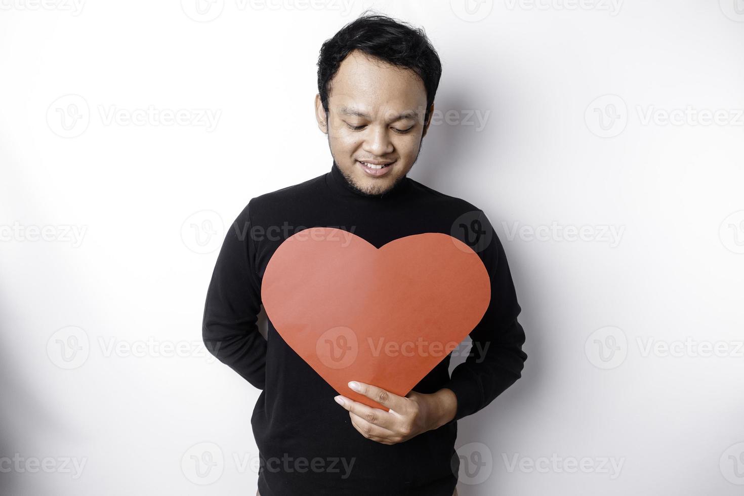 un' ritratto di un' contento asiatico uomo indossare un' nero camicia, Tenere un' rosso a forma di cuore carta isolato di bianca sfondo foto