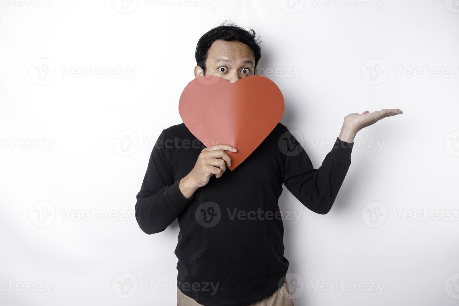 eccitato asiatico uomo indossare nero camicia, puntamento a il copia spazio accanto lui mentre Tenere un' grande rosso a forma di cuore carta, isolato di bianca sfondo foto