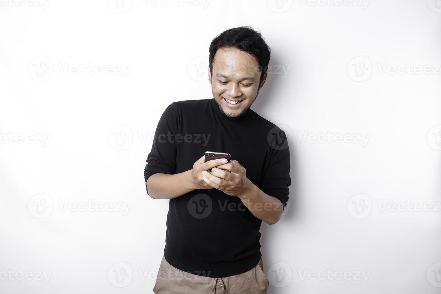 eccitato asiatico uomo indossare nero camicia sorridente mentre Tenere il suo Telefono, isolato di bianca sfondo foto