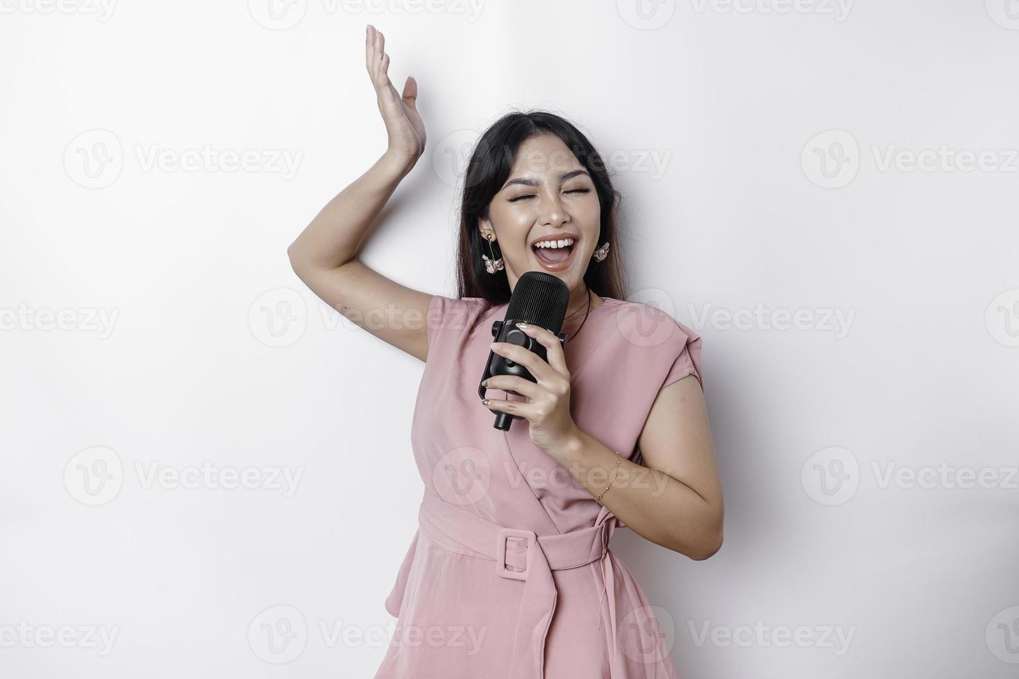 ritratto di spensierato asiatico donna, avendo divertimento karaoke, cantando nel microfono mentre in piedi al di sopra di bianca sfondo foto