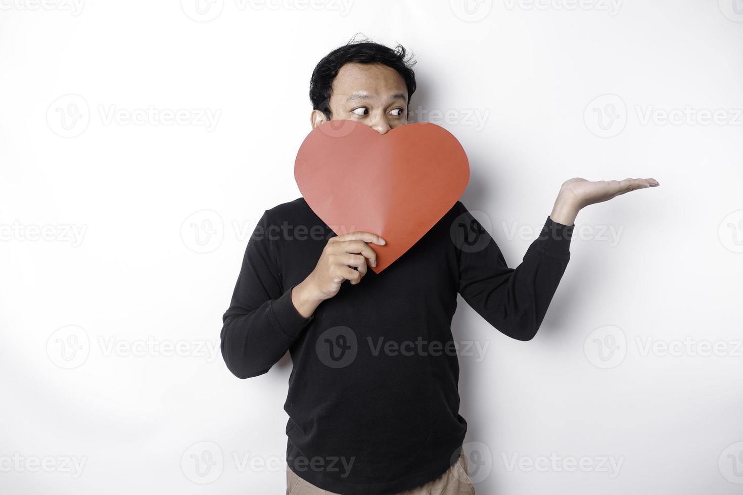 eccitato asiatico uomo indossare nero camicia, puntamento a il copia spazio accanto lui mentre Tenere un' grande rosso a forma di cuore carta, isolato di bianca sfondo foto