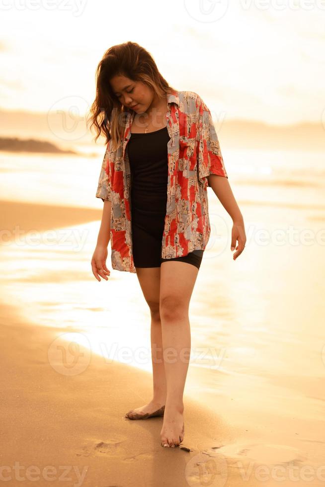 un asiatico donna con bionda capelli e un' rosso camicia passeggiate lungo il spiaggia per godere il mare Visualizza foto