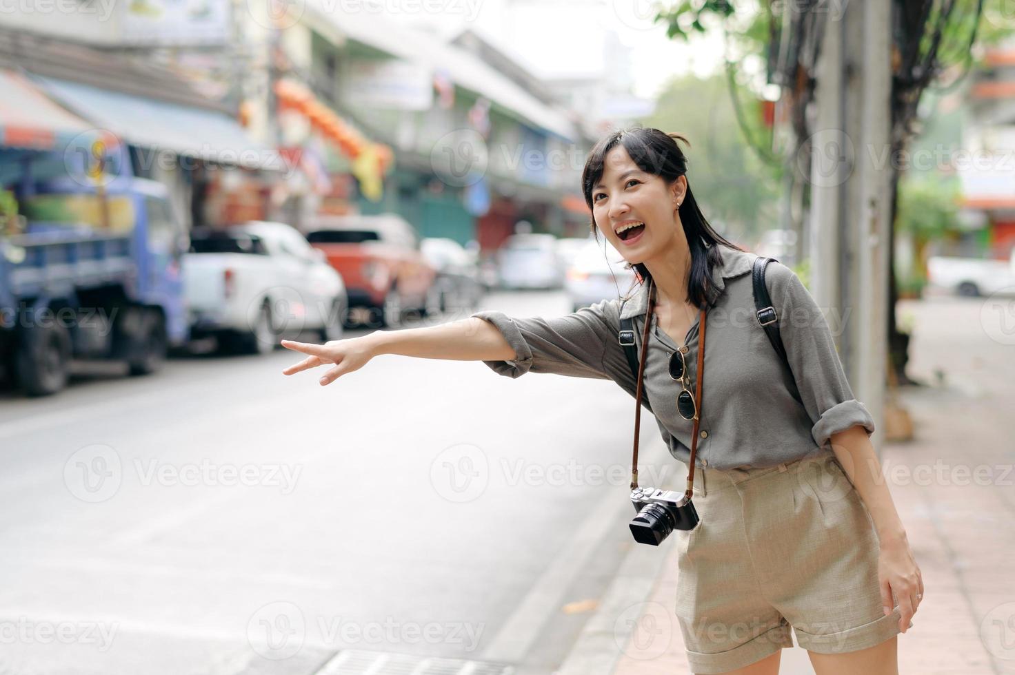 sorridente giovane asiatico donna viaggiatore autostop su un' strada nel il città. vita è un' viaggio concetto. foto