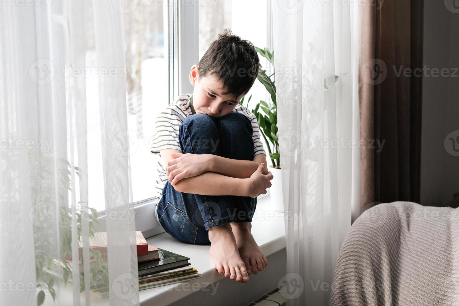 un' triste ragazzo si siede su il davanzale abbracciare il suo ginocchia. cattivo umore, depressione. il ragazzo è triste solo a casa foto