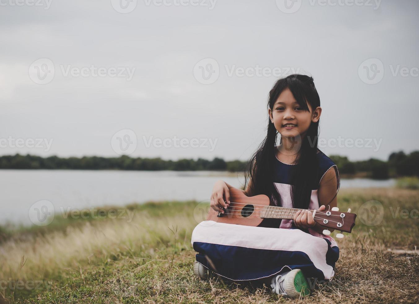 piccola ragazza asiatica sveglia che si siede sull'erba e suona l'ukulele nel parco foto