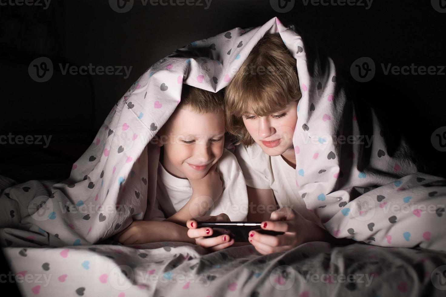 fratello e sorella menzogna sotto un' coperta a notte e giocare su il Telefono foto